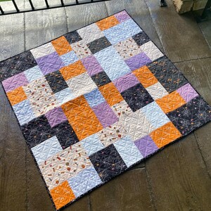 May include: A patchwork quilt with a Halloween theme. The quilt is made of various fabrics in orange, purple, black, and white. The quilt is quilted with a diamond pattern.