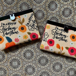 May include: Two small, floral patterned fabric wallets with black metal frames. The wallets have the text "I'm a ray of fucking sunshine" printed on them.