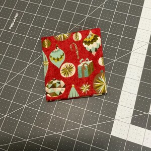 May include: A square piece of red fabric with a repeating pattern of Christmas ornaments in teal, gold, and white. The fabric also features images of wrapped gifts and candy canes. The fabric is placed on a gray cutting mat.