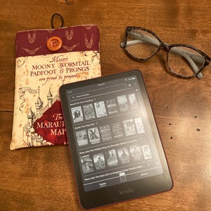 May include: An e-reader with a black screen displaying book covers, next to a book sleeve with text and a button closure, and a pair of tortoiseshell glasses. The book sleeve has the text "Messrs Moony, Wormtail, Padfoot & Prongs are proud to present The Marauder's Map."