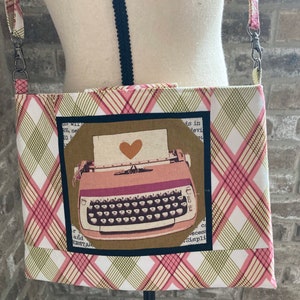 May include: A pink and green plaid crossbody bag with a vintage typewriter design. The typewriter has a heart-shaped paper holder and is surrounded by a black border.