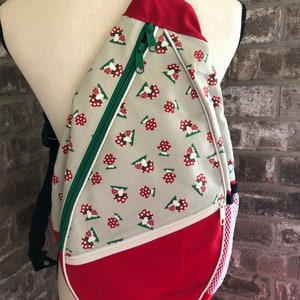 May include: A red and white sling bag with a mushroom pattern. The bag has a green zipper and a white mesh pocket on the side.