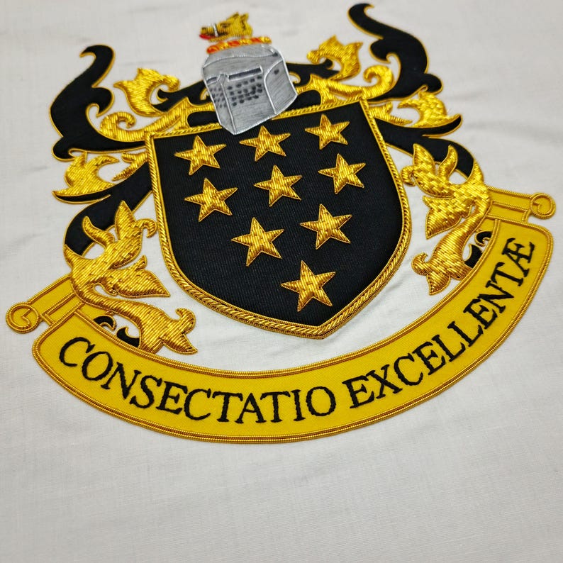 May include: Embroidered crest with a black shield featuring nine gold stars, a silver helmet, and gold filigree. The banner below reads "CONSECTATIO EXCELLENTE" in black lettering on a yellow background. The design is on a white fabric.