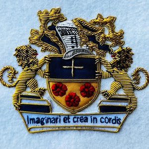 以下が含まれることがあります： 刺繍された紋章。十字架、3つの赤い花、ラテン語のテキスト「Imaginari et crea in cordis」が描かれた盾。2匹の金色のライオンが盾を挟み、金と銀の装飾が施されています。