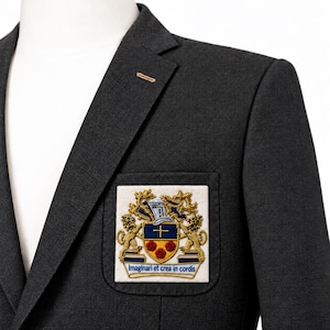 Puede incluir: Un blazer gris oscuro con solapa de muesca y un bolsillo en el pecho adornado con un parche rectangular. El parche muestra un escudo de armas bordado con leones dorados, un escudo azul con una cruz, flores rojas y el texto Imaginari et crea in cordis.