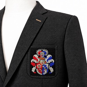 Puede incluir: Una americana de color gris carbón oscuro con un bolsillo de parche rectangular en el pecho. El bolsillo muestra un escudo bordado con hilos rojos, azules, blancos y plateados, que representa un yelmo y símbolos heráldicos.