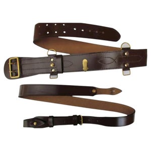 May include: A dark brown leather belt featuring a double-prong brass buckle, multiple adjustment holes, and brass hardware including D-rings and a central hook. The belt has reinforced stitching and a smooth finish, designed for utility and support.