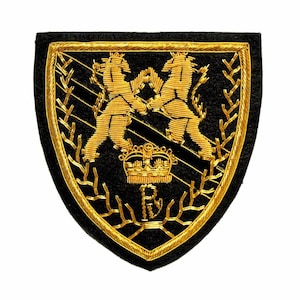 Puede incluir: Parche negro en forma de escudo con bordado dorado. Dos leones dorados se enfrentan sobre una corona dorada. El parche está enmarcado por un borde dorado y hojas de laurel doradas.