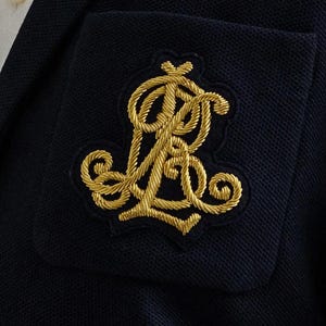 以下が含まれることがあります： 金色の刺繍モノグラムが付いた、濃紺のブレザーポケットのクローズアップ。モノグラムは、絡み合った文字を使った装飾的なデザインです。ブレザーは、テクスチャード加工された生地です。