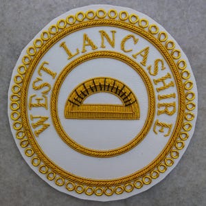 Op de afbeelding: Een wit embleem met gouden borduursel. De tekst "WEST LANCASHIRE" is gebogen rond een centrale cirkel. Binnen de cirkel bevindt zich een gouden geborduurde gradenboog. De buitenrand is afgewerkt met gouden touw en kleine gouden cirkels.