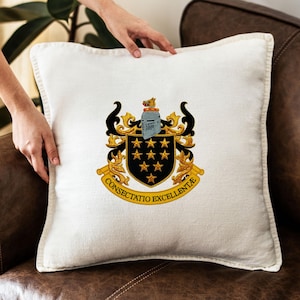 May include: A square white cushion featuring a detailed crest design. The crest includes a black shield with gold stars, a knight's helmet, and the words "CONSECTATIO EXCELLENTIAE" in gold. The cushion is on a brown leather sofa.