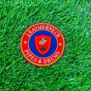 May include: A circular embroidered patch featuring a red border with a gold rope trim. The center is blue with a red shield containing a gold eagle, globe, and anchor emblem above a green shamrock. The text LEATHERNECK PIPES & DRUMS is in white.