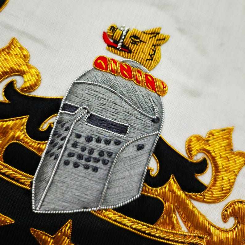May include: Detailed embroidery showing a knight's helmet, a golden lion, and gold filigree on a white backdrop. The helmet is grey with black detailing, and the lion has red accents. The design is likely part of a larger emblem or crest, possibly for heraldry.
