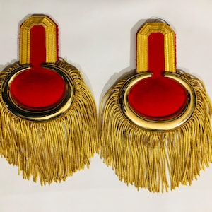 Puede incluir: Par de charreteras ornamentadas con centro de terciopelo rojo y flecos dorados. La sección superior es rectangular con ribete dorado, y la inferior presenta un diseño circular con borde dorado y largos flecos dorados.