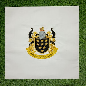 May include: White square cushion with an embroidered crest. The crest features a black shield with gold stars, a knight's helmet, and the words "CONSECTATIO EXCELLENTIA" in gold. The cushion is on a green surface.