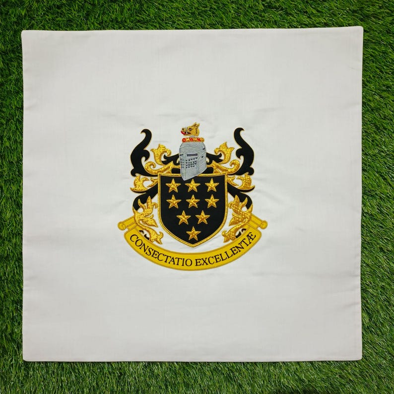 May include: White square cushion with an embroidered crest. The crest features a black shield with gold stars, a knight's helmet, and the words "CONSECTATIO EXCELLENTIA" in gold. The cushion is on a green surface.