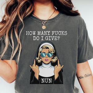 May include: Dark gray t-shirt with the text "HOW MANY FUCKS DO I GIVE?" above an illustration of a nun wearing sunglasses and giving the middle finger. The word "NUN" is below the illustration.