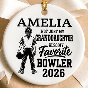 May include: A round white ceramic ornament with a gold hanging string. The design features a black graphic of a person holding a bowling ball and the text: AMELIA NOT JUST MY GRANDDAUGHTER ALSO MY FAVORITE BOWLER 2026 in black lettering.