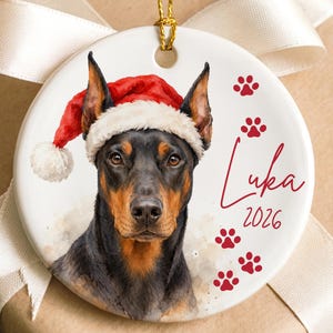 May include: A round white ceramic ornament featuring a Doberman Pinscher wearing a red and white Santa hat. The design includes the name Luka in red script, the year 2026, and several red paw prints. A gold string is attached for hanging.