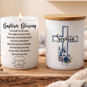 May include: Two white frosted glass candle jars. One is lit and features the text Baptism Blessing, a poem, and the date 12.20.2026 inside a heart. The other jar has a wooden lid and displays a blue cross with the name Sophia and floral accents.
