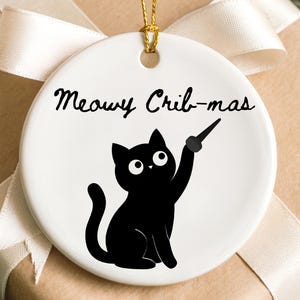 May include: A round white ceramic ornament featuring a black cat illustration holding a wand. The text "Meowy Crib-mas" is printed in black script above the cat. The ornament includes a gold string for hanging and is centered on a gift box.