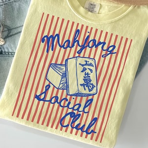 May include: A light yellow t-shirt with a red and white striped design and the words "Mahjong Social Club" in blue. The shirt is laid on top of a pair of light blue jeans and a tan hat.