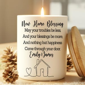 Puede incluir: Una vela blanca encendida con tapa de madera, con el texto "New Home Blessing" y una ilustraci&oacute;n de una casa. Se ven pi&ntilde;as en primer plano. El texto incluye los nombres "Emily & James".