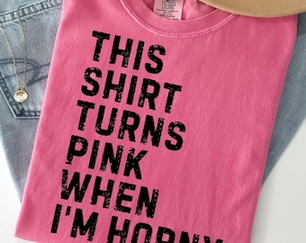 May include: A pink t-shirt with the text "THIS SHIRT TURNS PINK WHEN I'M HORNY" in black block letters. The shirt is folded and rests on a pair of light blue jeans. A tan hat is partially visible.