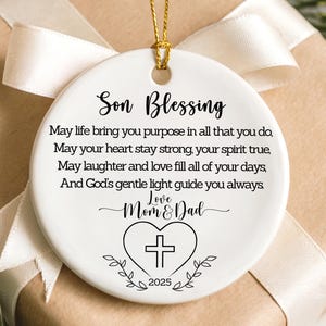 May include: A white ceramic ornament with a gold string for hanging. The ornament has the words "Son Blessing" and a heartfelt message printed on it, along with a cross inside a heart and the year 2025. The ornament is on a wrapped gift.