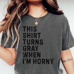 May include: Dark gray t-shirt with the text "THIS SHIRT TURNS GRAY WHEN I'M HORNY" in black. The shirt is a crew neck style and is worn with blue jeans. A gold necklace is visible.