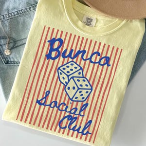 May include: A pale yellow t-shirt featuring a graphic print of two dice and the blue text "Bunco Social Club" centered over a background of vertical red stripes. The shirt has a crew neckline and a Comfort Colors brand label at the inner collar.