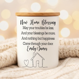 Puede incluir: Un tarro de vela de cer&aacute;mica blanca con tapa de madera. El tarro presenta el texto "New Home Blessing" y un mensaje personalizado, junto con un dibujo lineal de una casa y un coraz&oacute;n. Un regalo de inauguraci&oacute;n.
