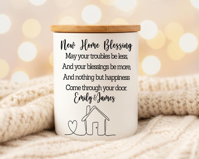 Puede incluir: Un tarro de vela de cer&aacute;mica blanca con tapa de madera. El tarro presenta el texto "New Home Blessing" y un mensaje personalizado, junto con un dibujo lineal de una casa y un coraz&oacute;n. Un regalo de inauguraci&oacute;n.