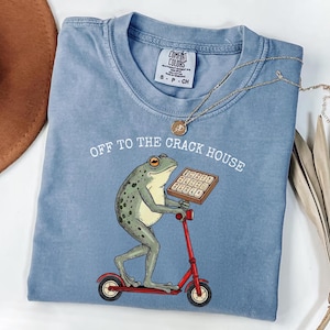 May include: A light blue Comfort Colors t-shirt with the phrase "OFF TO THE CRACK HOUSE" printed on it. The shirt features a cartoon frog riding a red scooter and holding a box. A gold necklace is draped across the shirt.