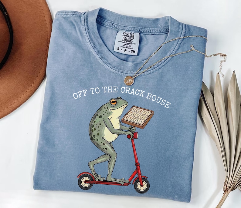 May include: A light blue Comfort Colors t-shirt with the phrase "OFF TO THE CRACK HOUSE" printed on it. The shirt features a cartoon frog riding a red scooter and holding a box. A gold necklace is draped across the shirt.