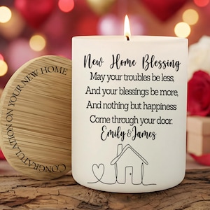 Puede incluir: Una vela blanca encendida con el texto "New Home Blessing" y un dibujo de una casa. Una posavasos de madera con las palabras "Felicitaciones por tu nuevo hogar" est&aacute; al lado de la vela. El fondo est&aacute; borroso con rojo y dorado.