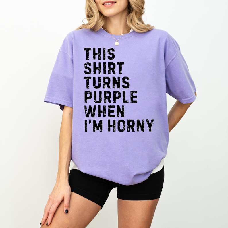 May include: A light purple t-shirt with the text "THIS SHIRT TURNS PURPLE WHEN I'M HORNY" in black block letters. The shirt is worn with black shorts.