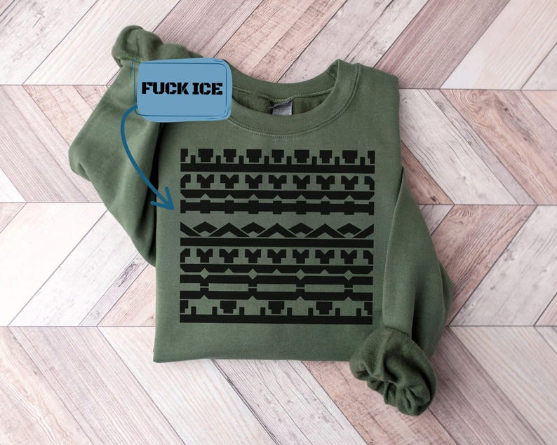 May include: An olive green sweatshirt with a black geometric pattern across the front. The text "FUCK ICE" is in a blue rectangle in the upper left corner. The sweatshirt is displayed on a light wood surface.