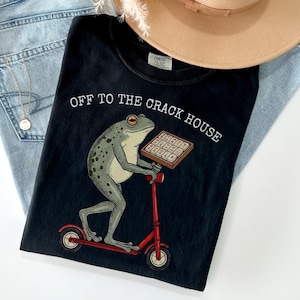May include: Black t-shirt with the text "OFF TO THE CRACK HOUSE" and a graphic of a frog riding a red scooter while holding a tray of waffles. The shirt is laid on top of a pair of blue jeans and a tan hat.