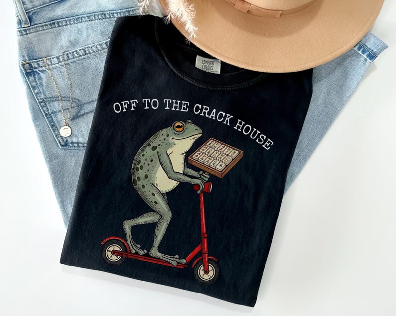 May include: Black t-shirt with the text "OFF TO THE CRACK HOUSE" and a graphic of a frog riding a red scooter while holding a tray of waffles. The shirt is laid on top of a pair of blue jeans and a tan hat.
