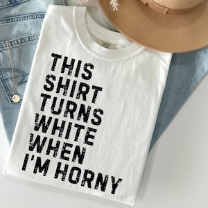 May include: White t-shirt with the text "THIS SHIRT TURNS WHITE WHEN I'M HORNY" in black block letters. The shirt is folded and rests on a pair of blue jeans. A tan hat is visible in the background.