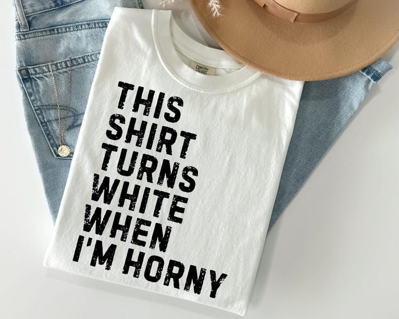 May include: White t-shirt with the text "THIS SHIRT TURNS WHITE WHEN I'M HORNY" in black block letters. The shirt is folded and rests on a pair of blue jeans. A tan hat is visible in the background.