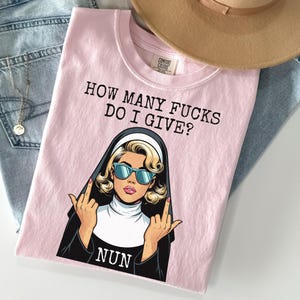 May include: A light pink t-shirt with the text "HOW MANY FUCKS DO I GIVE?" and a graphic of a nun wearing sunglasses and giving the middle finger. The shirt is folded on top of a pair of blue jeans and a tan hat.