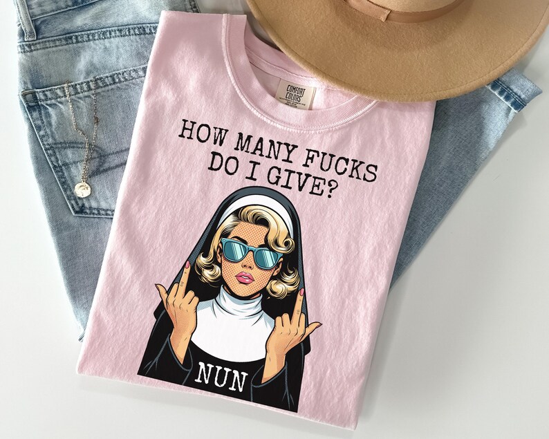 May include: A light pink t-shirt with the text "HOW MANY FUCKS DO I GIVE?" and a graphic of a nun wearing sunglasses and giving the middle finger. The shirt is folded on top of a pair of blue jeans and a tan hat.