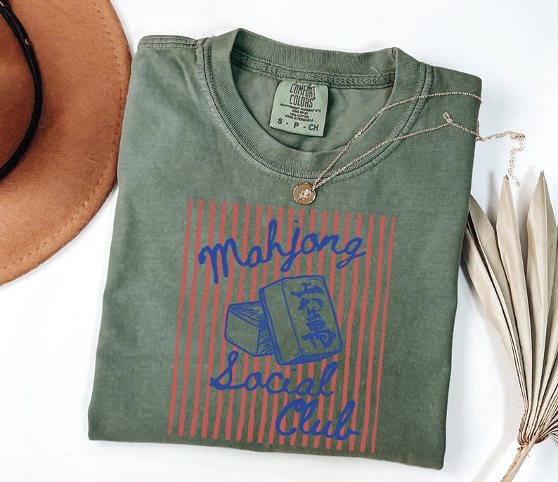 May include: Olive green t-shirt with a graphic that reads "Mahjong Social Club" in blue script, with red vertical stripes and a blue illustration of mahjong tiles. A gold necklace rests on the shirt. A brown hat is in the upper left corner.