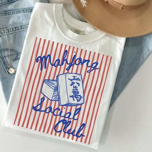 May include: White t-shirt with a red and white striped design and the words "Mahjong Social Club" in blue script. The shirt features a graphic of two Mahjong tiles. The shirt is laid on top of blue jeans.