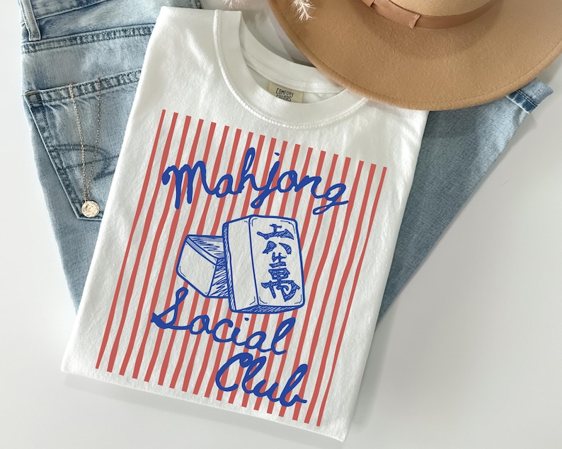 May include: White t-shirt with a red and white striped design and the words "Mahjong Social Club" in blue script. The shirt features a graphic of two Mahjong tiles. The shirt is laid on top of blue jeans.