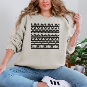 May include: A beige sweatshirt featuring a black geometric pattern across the front. The pattern consists of various shapes and lines arranged in horizontal rows. The sweatshirt has long sleeves and a crew neck.