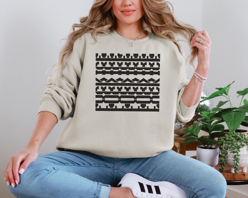 May include: A beige sweatshirt featuring a black geometric pattern across the front. The pattern consists of various shapes and lines arranged in horizontal rows. The sweatshirt has long sleeves and a crew neck.