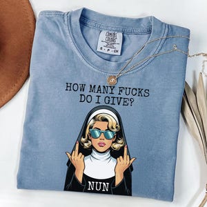 May include: A light blue t-shirt with the text "HOW MANY FUCKS DO I GIVE?" and a graphic of a nun wearing sunglasses and giving the middle finger. A gold necklace with a pendant is draped across the shirt.